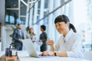 Professional woman in office setting multitasks when drinking water, colleagues in background